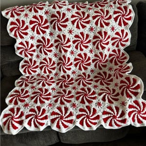 Może przedstawiać: Ręcznie robiony koc szydełkowy z powtarzającym się wzorem czerwono-białych wzorów z wirami miętowych cukierków. Koc ma falisty brzeg i jest narzucony na szarą sofę. Koc to przytulny, dekoracyjny przedmiot.