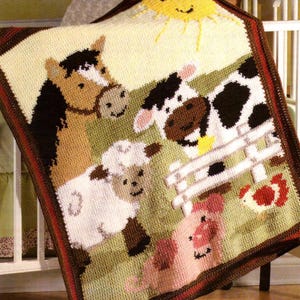 Op de afbeelding: Een gehaakte babydeken met een boerderijthema. De deken toont een paard, een koe, een schaap, een varken en een rode vogel. De achtergrond is een groen veld met een bruin hek.