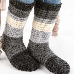 May include: A pair of handmade, knitted socks in shades of gray, with a cream-colored stripe. The socks are thick and appear warm, with a textured knit pattern. The socks are worn with blue jeans.