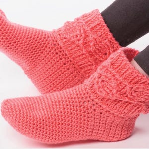 May include: Coral-coloured knitted slipper boots. The boots have a textured, folded cuff and a rounded toe. The thick knit construction suggests warmth and comfort. The boots are shown on a white background.