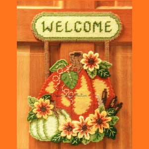 May include: A handmade fall-themed welcome sign. The sign features a pumpkin and sunflowers, with the word "WELCOME" stitched across the top. The pumpkin is orange and yellow, with green leaves and a green pumpkin. The sunflowers are yellow and orange.