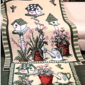 Könnte beinhalten: Eine cremefarbene, gestrickte Decke und ein passender Teppich mit einer Gartenszene. Das Design umfasst Vogelhäuser, rosa Blumen und eine Gießkanne. Der Rand ist grün und weiß gestreift. Der Text "Beesia Vintage Collection" ist sichtbar.