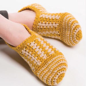 May include: A pair of handmade, yellow and beige crochet slippers. The slippers feature a checkered pattern on the top and a solid yellow trim. The cozy footwear is designed for indoor use and provides warmth and comfort.