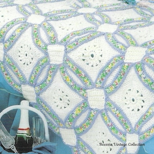 Puede incluir: Primer plano de una manta blanca de ganchillo con un patrón repetitivo de cuadrados y círculos entrelazados. Los círculos están adornados con un estampado floral y bordes azul claro. La manta está sobre una cama con una falda de cama azul. Una botella de champán y copas en primer plano.