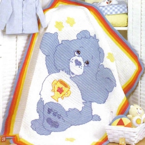 May include: A crocheted baby blanket featuring a blue Care Bear holding a gold trophy with a star on it. The blanket has a rainbow border and is made with white yarn.