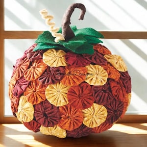 May include: A handmade, round pumpkin decoration with a stem and leaves. The pumpkin is covered in layered, fabric circles in shades of orange, yellow, and burgundy. The stem is brown, and the leaves are green. The pumpkin is a fall-themed decorative item.