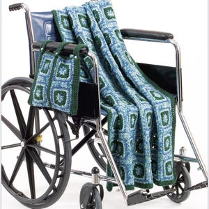 May include: A blue and green crocheted blanket and matching bag draped over a wheelchair. The blanket features a square pattern with green borders. The bag hangs from the armrest. The wheelchair has black wheels and a blue seat.