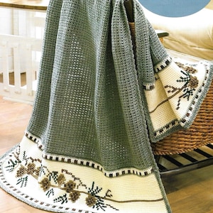 May include: An olive green crochet blanket with a detailed border. The border has a cream-coloured band with embroidered pine cone and branch designs in brown and green. The blanket is draped over a chair.