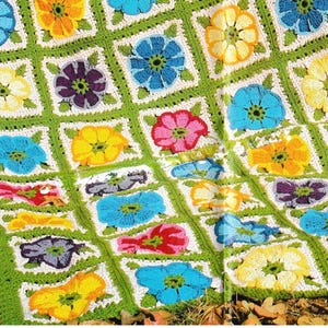 May include: A vibrant, hand-crocheted blanket with a floral design. The blanket displays squares of flowers in shades of blue, yellow, pink, and purple, set against a white background and a green border.