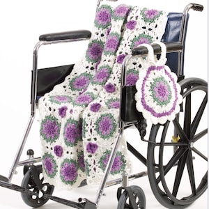 Puede incluir: Una manta y un posavasos redondos de ganchillo en una silla de ruedas. La manta presenta un fondo blanco con patrones florales morados y verdes. El posavasos tiene un diseño similar, con un fondo blanco y detalles morados y verdes.