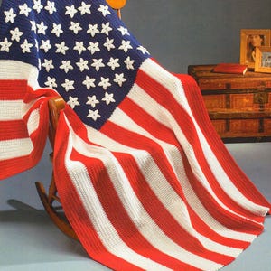 May include: A crocheted American flag blanket in red, white, and blue, draped over a rocking chair. The stars are made of white yarn and the stripes are made of red and white yarn.