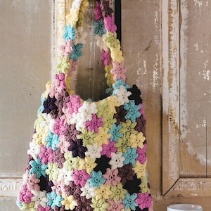 May include: A handmade crochet tote bag featuring a floral pattern in various colors, including pink, yellow, blue, and purple. The bag has a sturdy handle and is hanging against a rustic wooden door. The bag's dimensions are not visible.