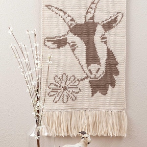 Könnte beinhalten: Ein gehäkelter Wandbehang mit einer braunen Ziege, die eine weiße Blume im Maul hält. Der Behang hat einen Fransenrand und ist an einer Holzleiste befestigt.