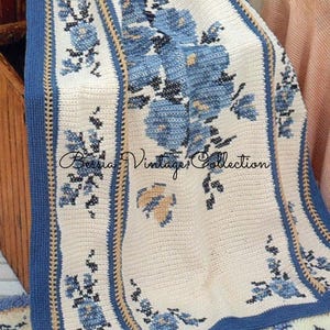 Może przedstawiać: Kremowy, szydełkowy koc z kwiatowym wzorem w odcieniach niebieskiego. Koc ma niebiesko-beżową obwódkę w paski, z napisem "Bessia Vintage Collection" w kursywie.
