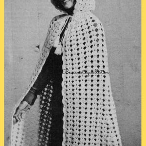 May include: A cream-coloured, open-weave crochet cape with a hood and tassel ties. The cape has a diamond pattern and a scalloped edge. The text "Bessiavintagetcollection" is visible on the cape. The cape is worn over a dark-coloured garment.