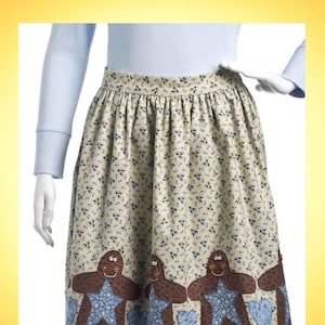 Puede incluir: Delantal con un estampado floral beige y un dobladillo festoneado. El delantal presenta una fila de aplicaciones de hombrecitos de jengibre con detalles de estrellas y corazones azules. El texto "Gingerbread Apron" está en la parte superior.