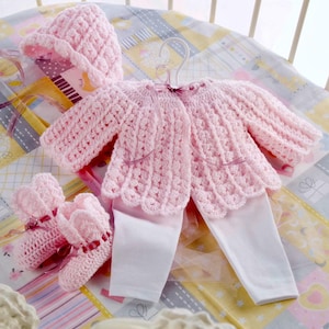 May include: A baby's crochet outfit set in soft pink, including a jacket, hat, leggings, and booties. The jacket and hat have scalloped edges and ribbon accents. The set is displayed on a patterned blanket.