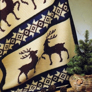Pode incluir: Um cobertor de crochê creme com um padrão repetido de silhuetas de renas marrons escuras e desenhos geométricos. O cobertor tem listras azul marinho com padrões geométricos. O cobertor está dobrado, com uma pequena árvore perene e pinhas em primeiro plano.