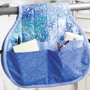 May include: A blue and white fabric bed rail organiser with multiple pockets. The top section has a floral pattern, while the pockets have a solid blue and striped design. It holds a notepad, glasses, and other small items. The organiser is designed to hang from a bed rail.