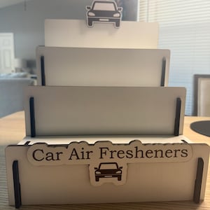 Könnte beinhalten: Ein mehrstufiger Präsentationsständer für Auto-Lufterfrischer. Der Ständer ist aus hellem Holz gefertigt und zeigt eine Autografik. Der Text "Car Air Fresheners" ist auf der Vorderseite zu sehen.