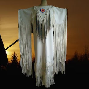 May include: A white dress with long fringe sleeves and hem. The dress features a V-neck with a circular emblem and beaded accents. The dress is displayed on a mannequin against a sunset backdrop.