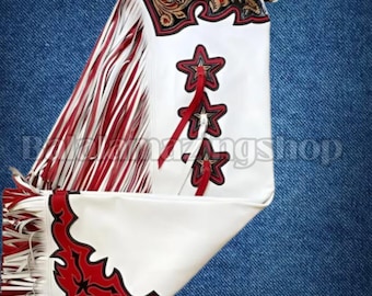 Könnte beinhalten: Weiße Lederchaps mit roten und schwarzen Zierakzenten. Zu den Merkmalen gehören rote Sterndesigns, Fransen und ein brauner Ledergürtel. Die Chaps haben eine dekorative schwarze und goldene Oberseite.