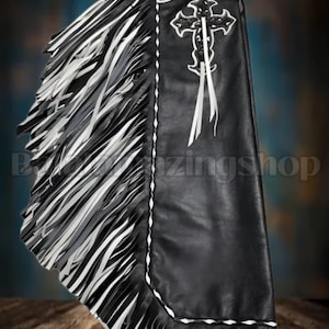 Puede incluir: Chaps de cuero negro con flecos blancos y grises. Los chaps presentan un diseño de cruz blanca con cintas blancas y costuras decorativas. La parte superior de los chaps tiene una correa negra.