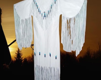 May include: White dress with long sleeves and fringe detailing. The dress features a V-neckline adorned with blue and silver beads and fringe. Additional blue and silver beads are placed vertically down the front of the dress. The sleeves and hemline are finished with long fringe.