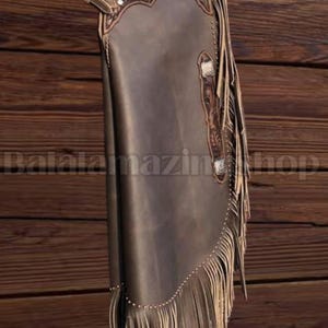 K&ouml;nnte beinhalten: Braune Lederchaps mit dekorativen Verzierungen und Fransen. Die Chaps weisen ein detailliertes Blumendesign im oberen Bereich auf und sind mit silberfarbener Hardware akzentuiert. Das Bild ist vor einem Holzgrund platziert.