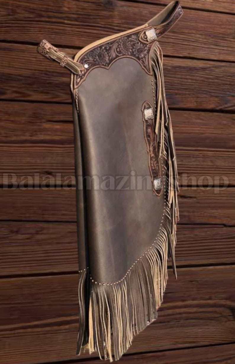 K&ouml;nnte beinhalten: Braune Lederchaps mit dekorativen Verzierungen und Fransen. Die Chaps weisen ein detailliertes Blumendesign im oberen Bereich auf und sind mit silberfarbener Hardware akzentuiert. Das Bild ist vor einem Holzgrund platziert.