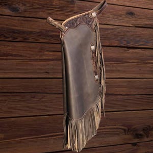 K&ouml;nnte beinhalten: Dunkelbraune Lederchaps mit dekorativen Verzierungen und Fransen. Die Chaps haben ein detailliertes Design im oberen Bereich und an den Seiten. Die Fransen sind hellbraun.