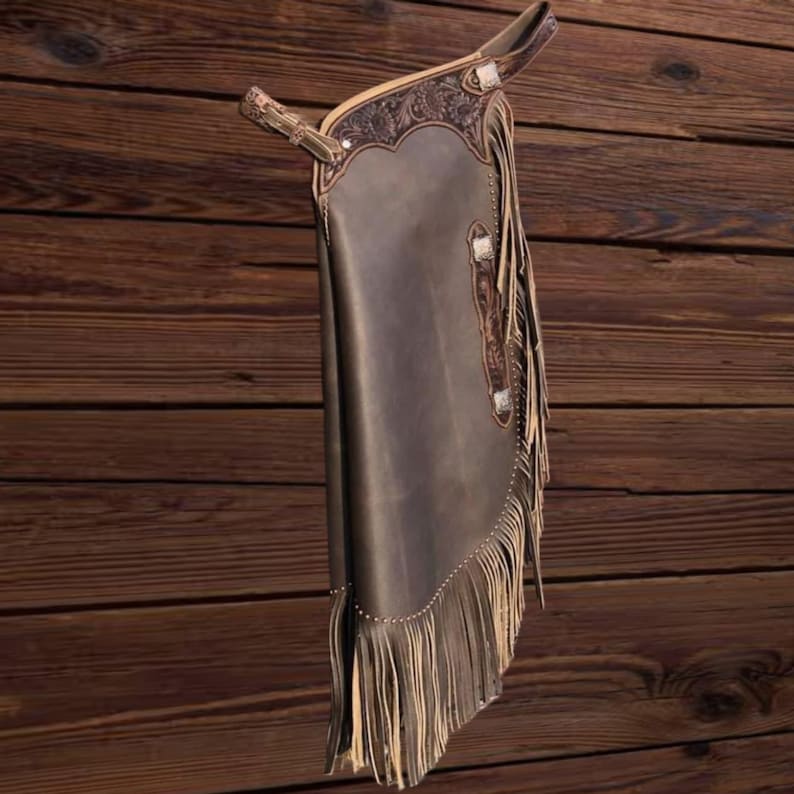 K&ouml;nnte beinhalten: Dunkelbraune Lederchaps mit dekorativen Verzierungen und Fransen. Die Chaps haben ein detailliertes Design im oberen Bereich und an den Seiten. Die Fransen sind hellbraun.