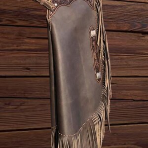 K&ouml;nnte beinhalten: Braune Lederchaps mit dekorativen Verzierungen und Fransen. Die Chaps weisen ein detailliertes Design im oberen Bereich und an den Kanten auf. Silberfarbene Beschl&auml;ge akzentuieren das Design. Die Chaps werden vor einem Holzgrund gezeigt.