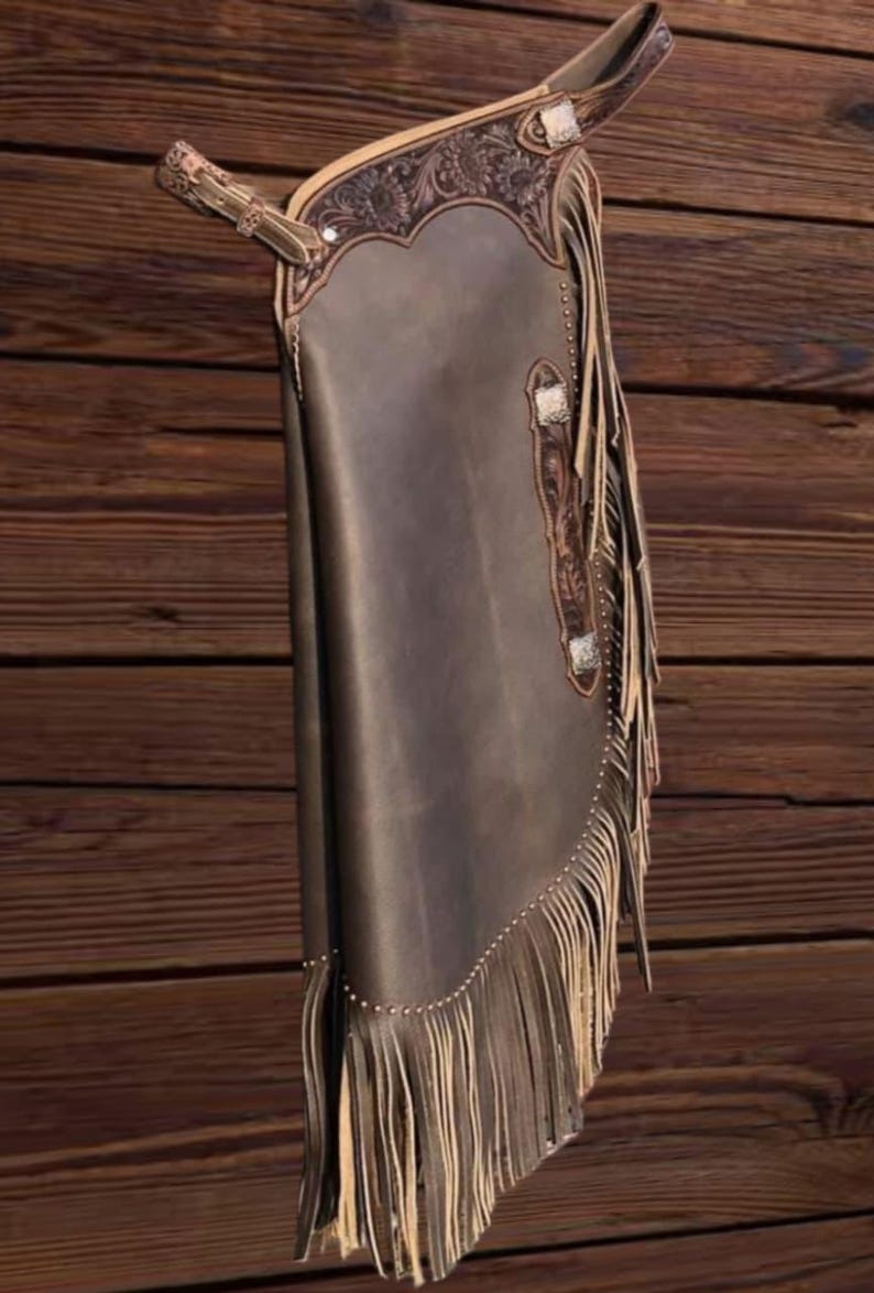 K&ouml;nnte beinhalten: Braune Lederchaps mit dekorativen Verzierungen und Fransen. Die Chaps weisen ein detailliertes Design im oberen Bereich und an den Kanten auf. Silberfarbene Beschl&auml;ge akzentuieren das Design. Die Chaps werden vor einem Holzgrund gezeigt.