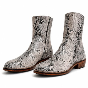 May include: A pair of ankle boots featuring a grey and black snakeskin pattern. The boots have a rounded toe, a side zipper closure, and a stacked wooden heel with a dark brown sole. The textured material covers the entire upper portion of the boots.