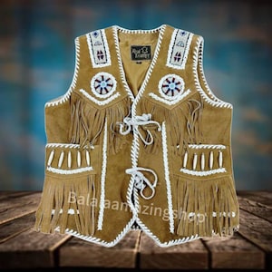 Puede incluir: Un chaleco de ante marrón con flecos y abalorios blancos. El chaleco tiene dos bolsillos con detalles de hueso y un diseño de abalorios alrededor del cuello. El chaleco está sobre una superficie de madera.