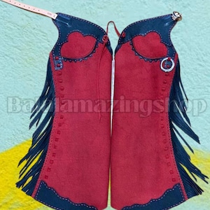 May include: Red suede chaps with black leather accents and fringe. The chaps feature a decorative heart-shaped design and are secured with a leather strap and metal hardware. The chaps are designed for riding.