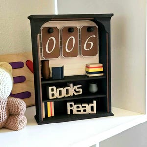 May include: A black wooden bookshelf with the numbers "006" displayed on brown panels. The shelf contains miniature books, a vase, and the words "Books Read" in wooden letters. The shelf is decorated with colorful accents.
