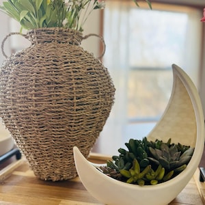 May include: A white ceramic crescent moon planter with a succulent plant inside. The planter is sitting on a wooden tray next to a woven wicker vase with yellow and pink flowers.