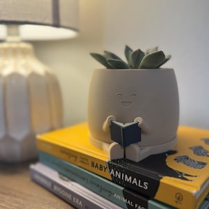 May include: A matte white 3d printed planter with a smiley face sitting with legs crossed and holding a navy blue book. The planter is sitting on top of a stack of three books.