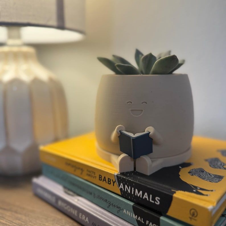May include: A matte white 3d printed planter with a smiley face sitting with legs crossed and holding a navy blue book. The planter is sitting on top of a stack of three books.