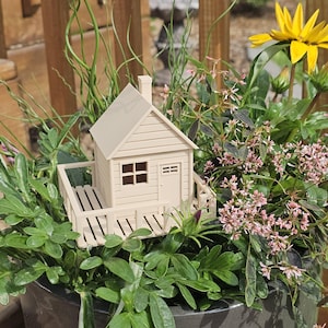 May include: A miniature beige house with a small porch and a chimney, nestled among green plants and pink flowers. The house has a door and a window with a cross design. The scene is set in a decorative planter.