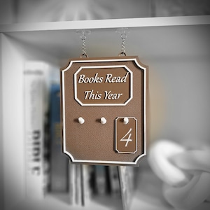 May include: A brown sign with white trim and the text "Books Read This Year." The sign has three small pegs and a tag with the number "4." The sign is hanging from a shelf.
