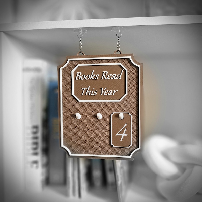May include: A brown sign with white trim and the text "Books Read This Year." The sign has three small pegs and a tag with the number "4." The sign is hanging from a shelf.