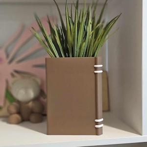 May include: A brown rectangular planter shaped like a book, with white accents on the spine, holding green grass. The planter is on a white shelf, with a blurred pink floral design and a teddy bear in the background.