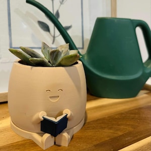 May include: A beige ceramic planter shaped like a person sitting with a book. The planter has a smiling face and is holding a black book. A succulent plant is growing in the planter. A green watering can is in the background.