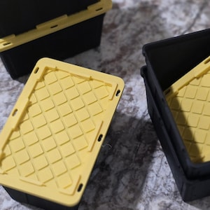 May include: Three black storage containers with yellow lids. The lids have a diamond pattern and are rectangular. The containers are made of plastic and are designed for storage. The lids are removable and fit snugly on the containers.