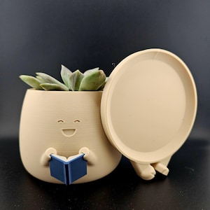May include: A beige 3d printed planter shaped like a smiling face with a blue book in its hands. The planter has a removable lid and a drainage hole. The planter is filled with a green succulent plant.