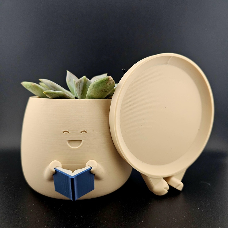 May include: A beige 3d printed planter shaped like a smiling face with a blue book in its hands. The planter has a removable lid and a drainage hole. The planter is filled with a green succulent plant.