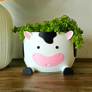 May include: A white cow-shaped planter with black spots, pink ears, and a pink mouth. The planter is filled with green plants. The planter has black feet and is sitting on a wooden surface.